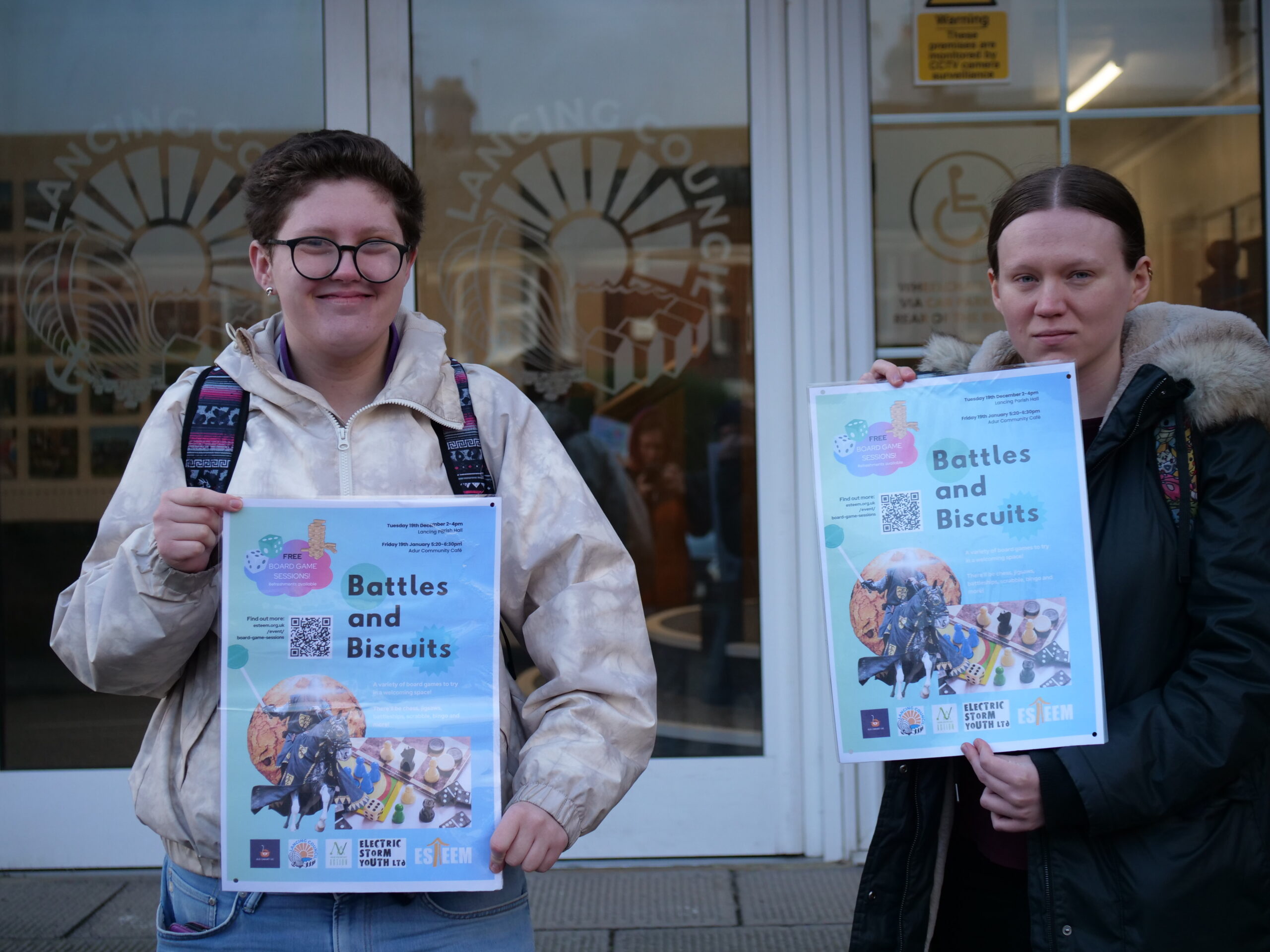 Two young adults smile holding posters for an intergenerational board game event